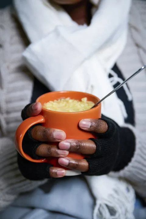 A close-up of a person’s gloved hands holding a mug of cup noodles.